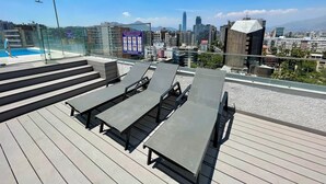 Una terraza en la azotea con tumbonas y piscina, con vistas a un paisaje urbano que incluye edificios modernos y montañas al fondo.