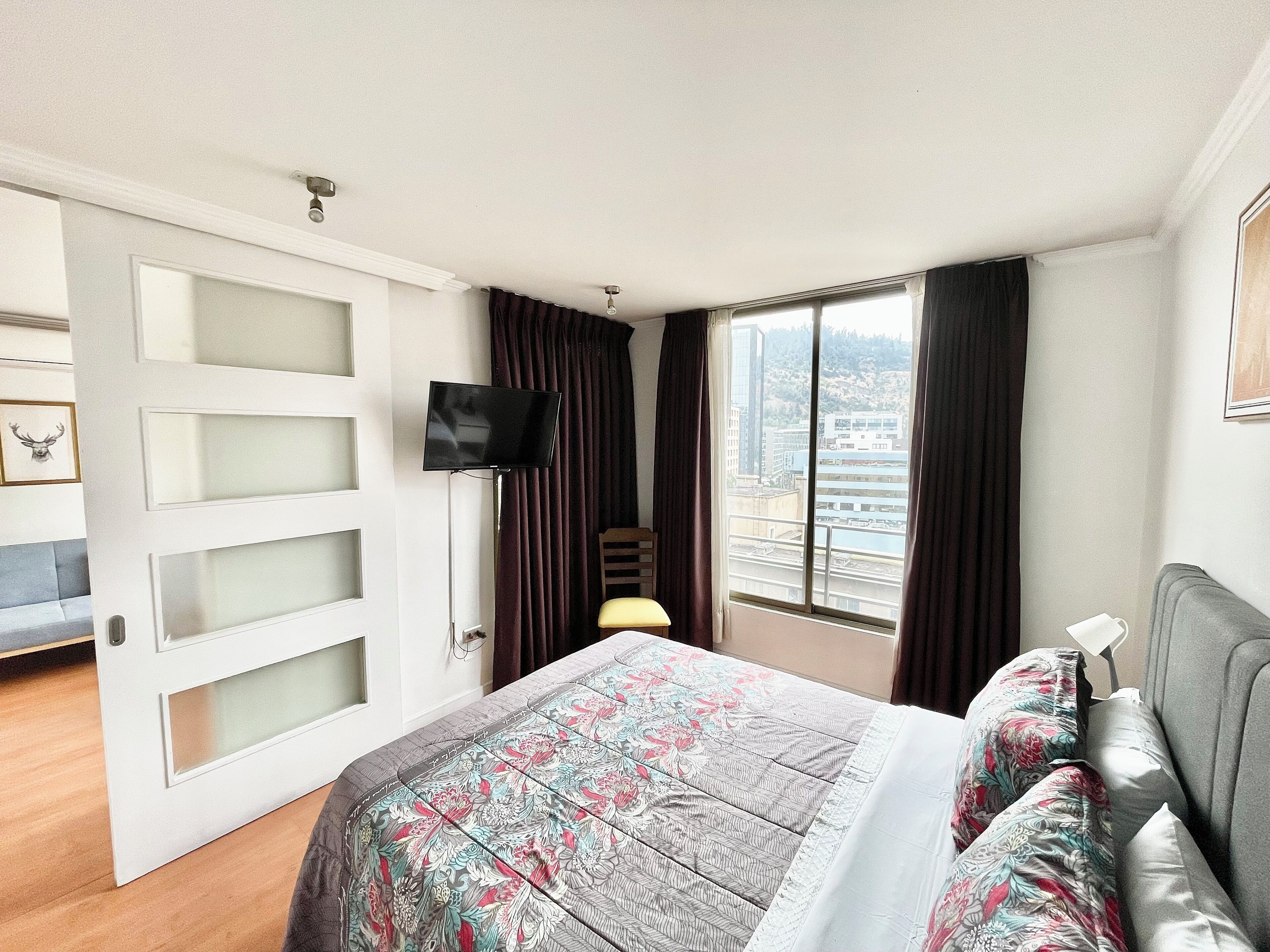 A bedroom with a bed, a TV, a window with curtains, and a city view.