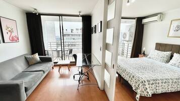 A modern hotel room with a grey sofa, a bed with patterned bedding, a small table with chairs, and a balcony with a view of the city.