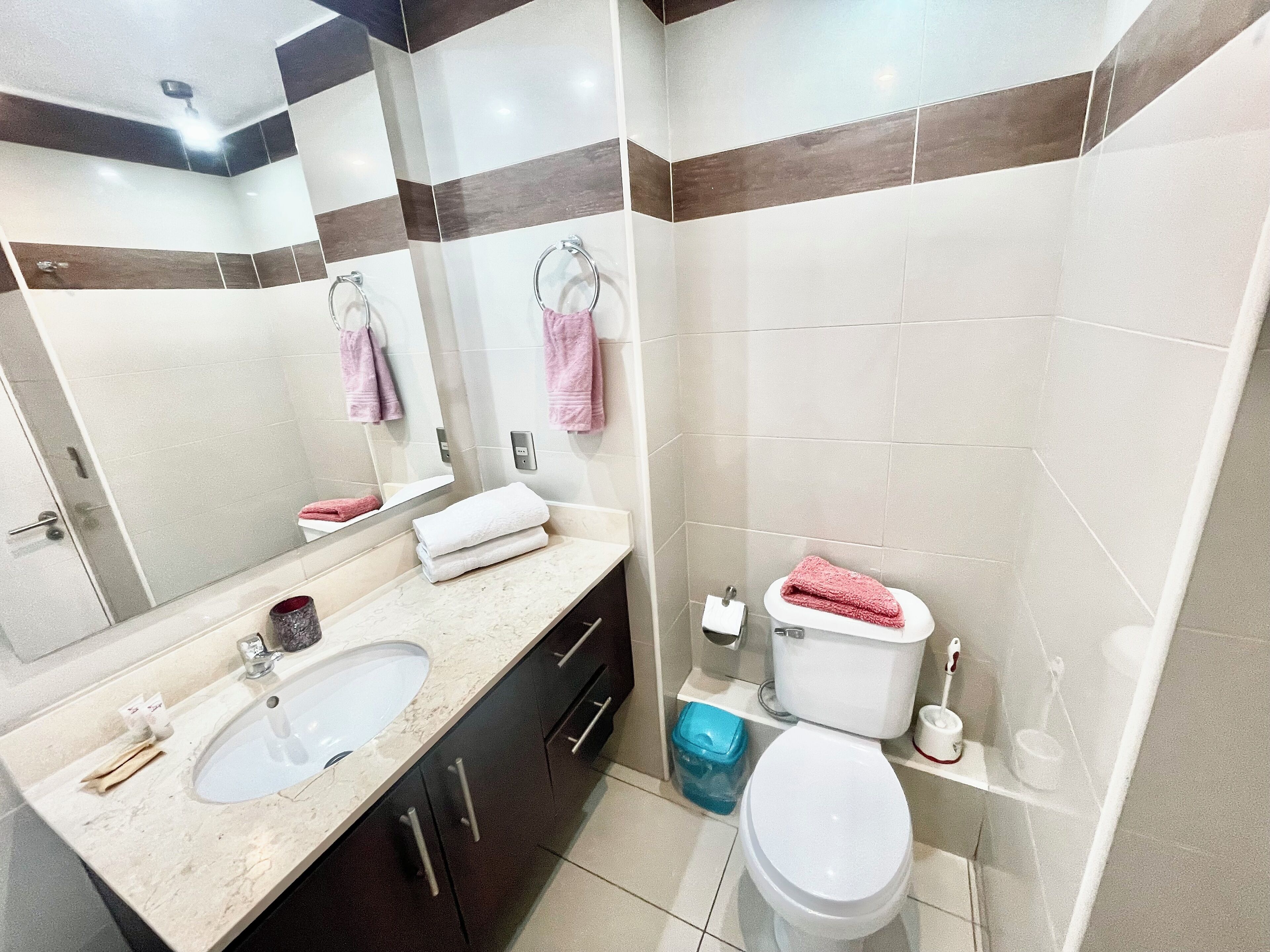 Un baño con dos lavamanos, un inodoro, un espejo y un cubo de basura.