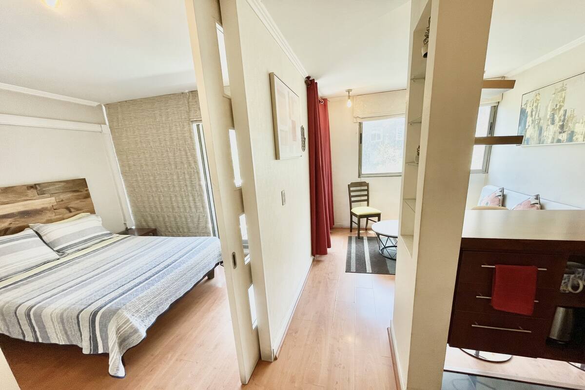 A hotel room with a bed, a desk, a chair, and a kitchen area.