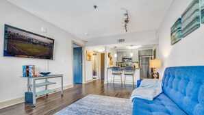 Un salon avec un canapé bleu, un téléviseur fixé au mur, une cuisine en arrière-plan et un coin repas avec des tabourets de bar.