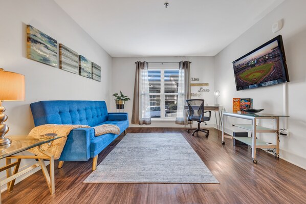 A modern living room with a blue sofa, a glass coffee table, a desk with a computer, and a large TV on the wall.