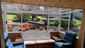 Een oranjerie met een gewatteerde bank, twee fauteuils en uitzicht op een tuin met rode en groene planten.