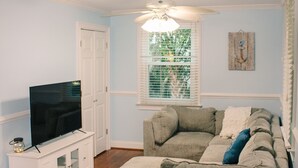 Ein Wohnzimmer mit einer Eckcouch, einem Fernseher auf einem Schrank, einem Deckenventilator und einem Fenster mit Jalousien.