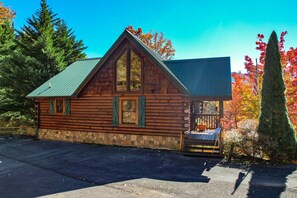 A log cabin with a stone foundation, a steeply pitched roof, and a porch with a railing.