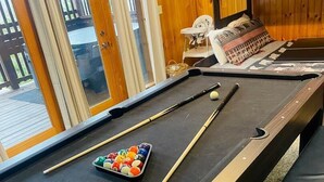 A pool table with balls and cues, a bench, and a bunk bed in a room with wooden walls and a glass door.