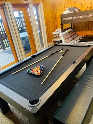Ein Billardtisch mit Kugeln und Spielstöcken, eine Bank und ein Etagenbett in einem Raum mit Holzwänden und einer Glastür.