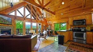 Interior espacioso de una cabaña de troncos con chimenea, comedor, cocina y ventanas amplias que ofrecen vistas panorámicas.