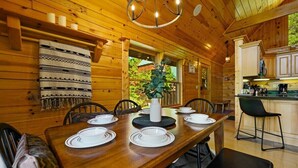 A wooden cabin interior with a dining table set for four, a kitchen area, and a large window with a view of greenery.