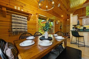 Interior de una cabaña de madera con una mesa de comedor preparada para cuatro personas, una zona de cocina y un amplio ventanal con vistas a la vegetación.