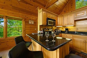 Une cuisine rustique aménagée dans une cabane en bois, avec un îlot central, des meubles en bois et une grande fenêtre donnant sur un paysage verdoyant.