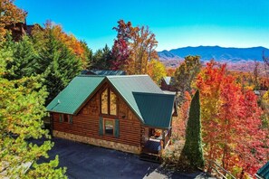 Une cabane en rondins surmontée d’un toit végétalisé, entourée d’un feuillage automnal et avec des montagnes en arrière-plan.