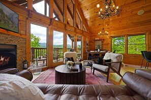A cozy log cabin interior with a fireplace, leather seating, a dining area, and large windows offering a view of the outdoors.