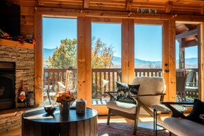 Interior de una cabaña con chimenea de piedra, mobiliario de madera y un ventanal con vistas a un paisaje montañoso.