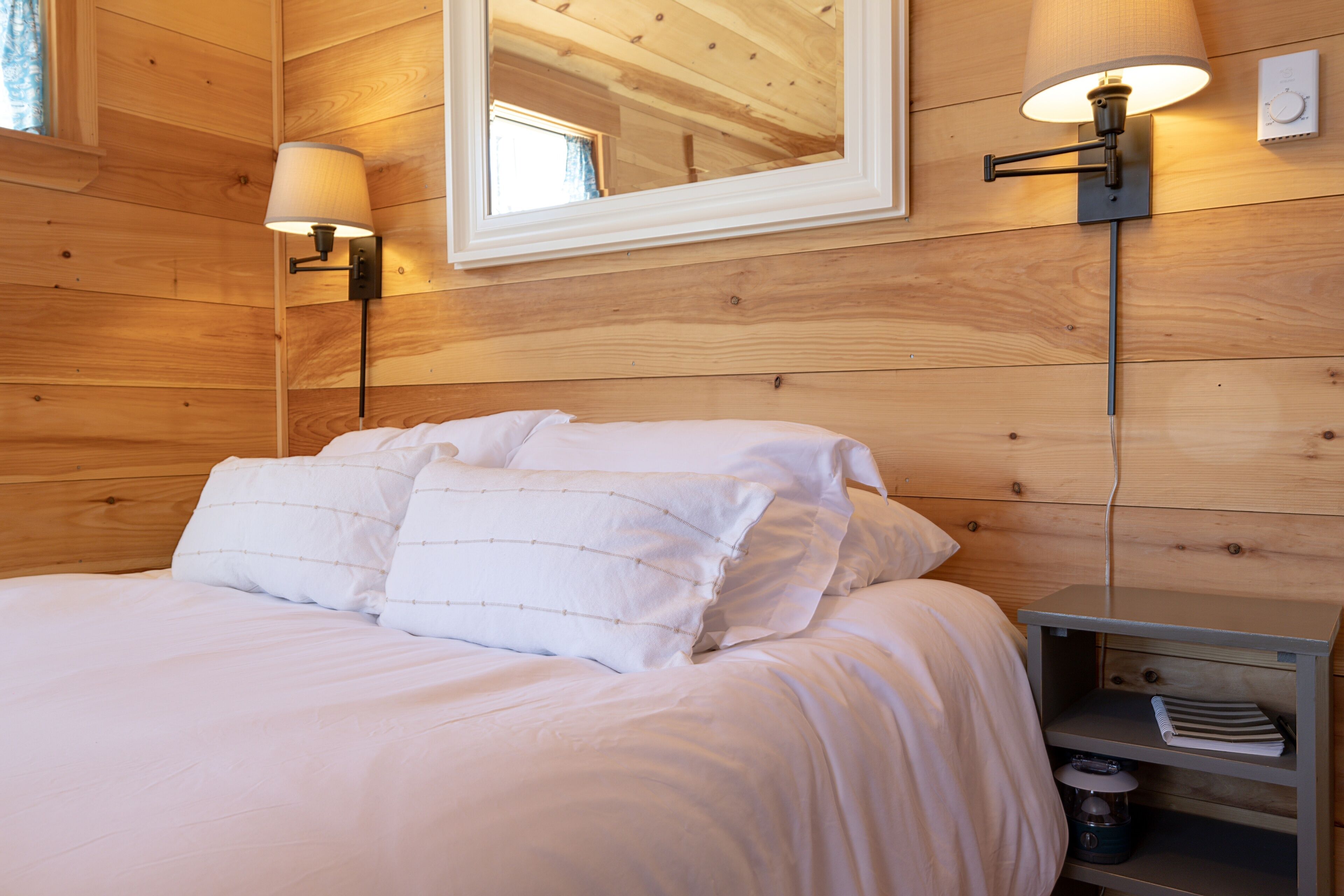 A wooden-paneled bedroom with a bed, two wall-mounted lamps, a mirror, and a nightstand with a lamp and a kettle.