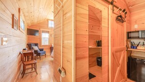 A wooden cabin interior with a dining area, a living room with a sofa and armchair, and a kitchen with a refrigerator and shelves. - Cabin 1 Near Sandy Point Beach + Wood-Fired Sauna (Stockton Springs)