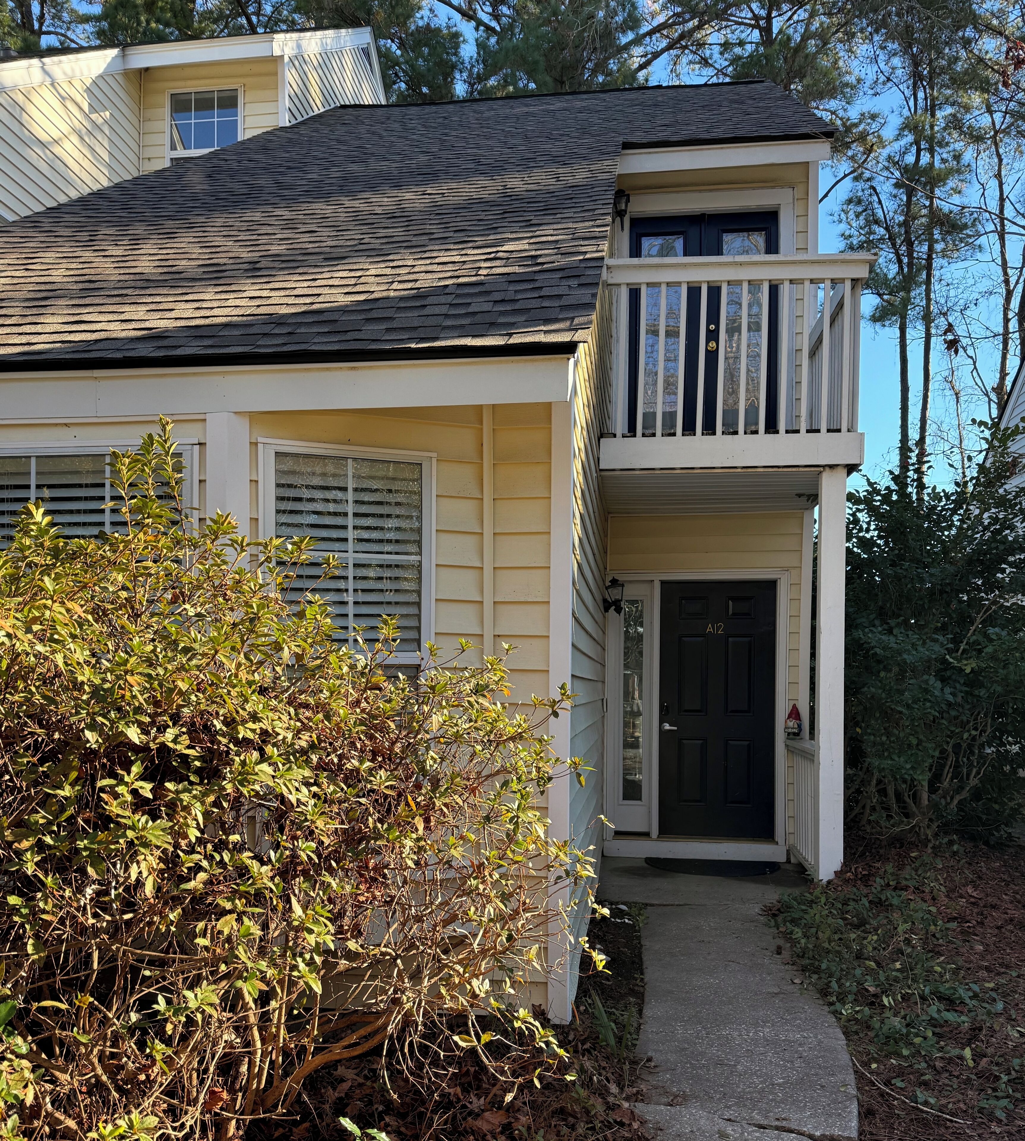 A two-story house with a balcony, a front door labeled "A12", and a small garden area with bushes and a pathway leading to the door.