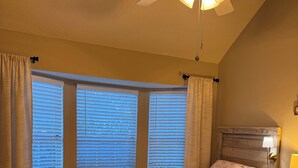 Ein Schlafzimmer mit einem Bett, einem Nachttisch, einer Lampe, einem Deckenventilator und einem Fenster mit Jalousien.