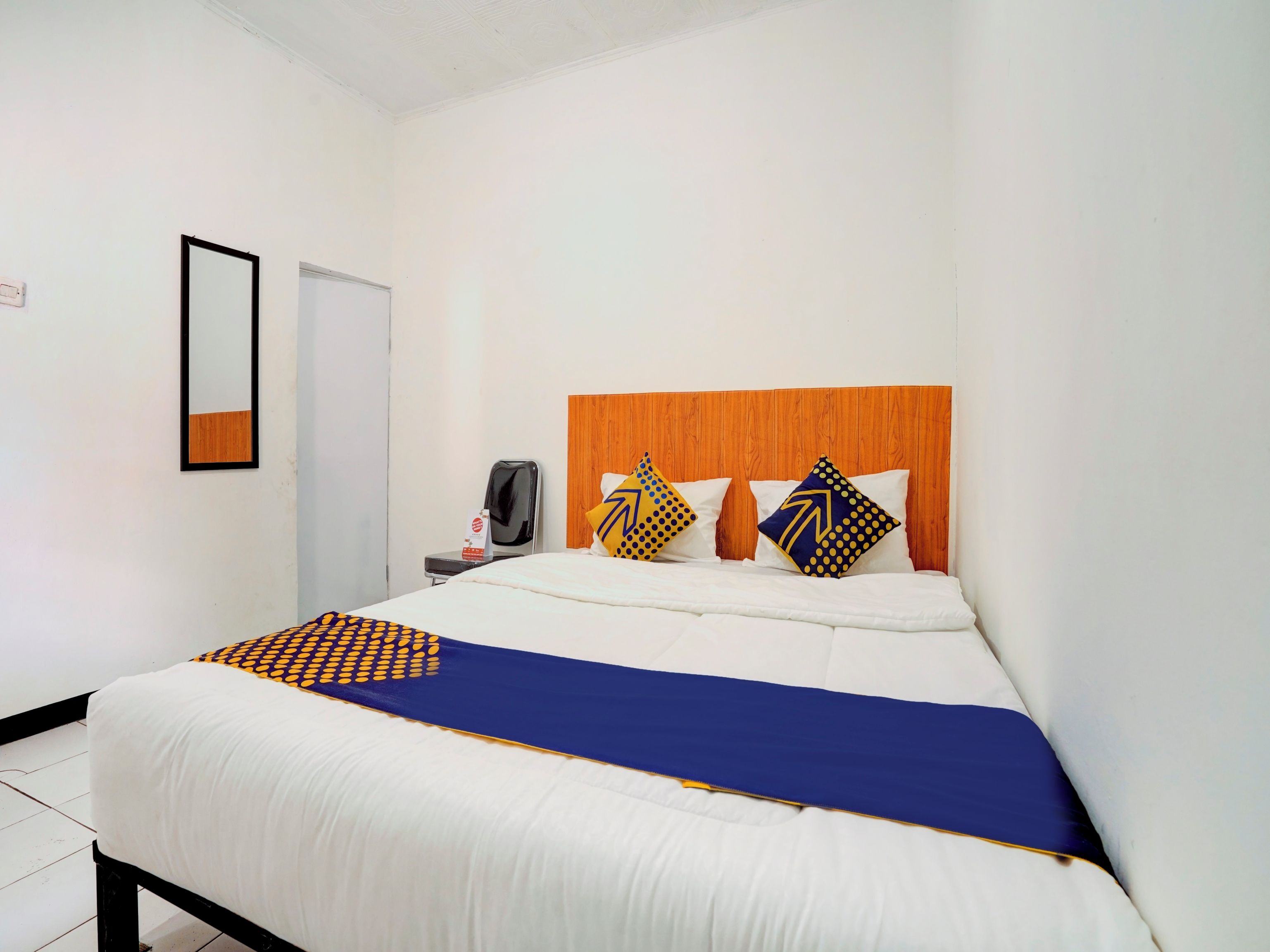 A hotel room with a bed, a wooden headboard, a desk with a chair, a mirror, and a wall socket.