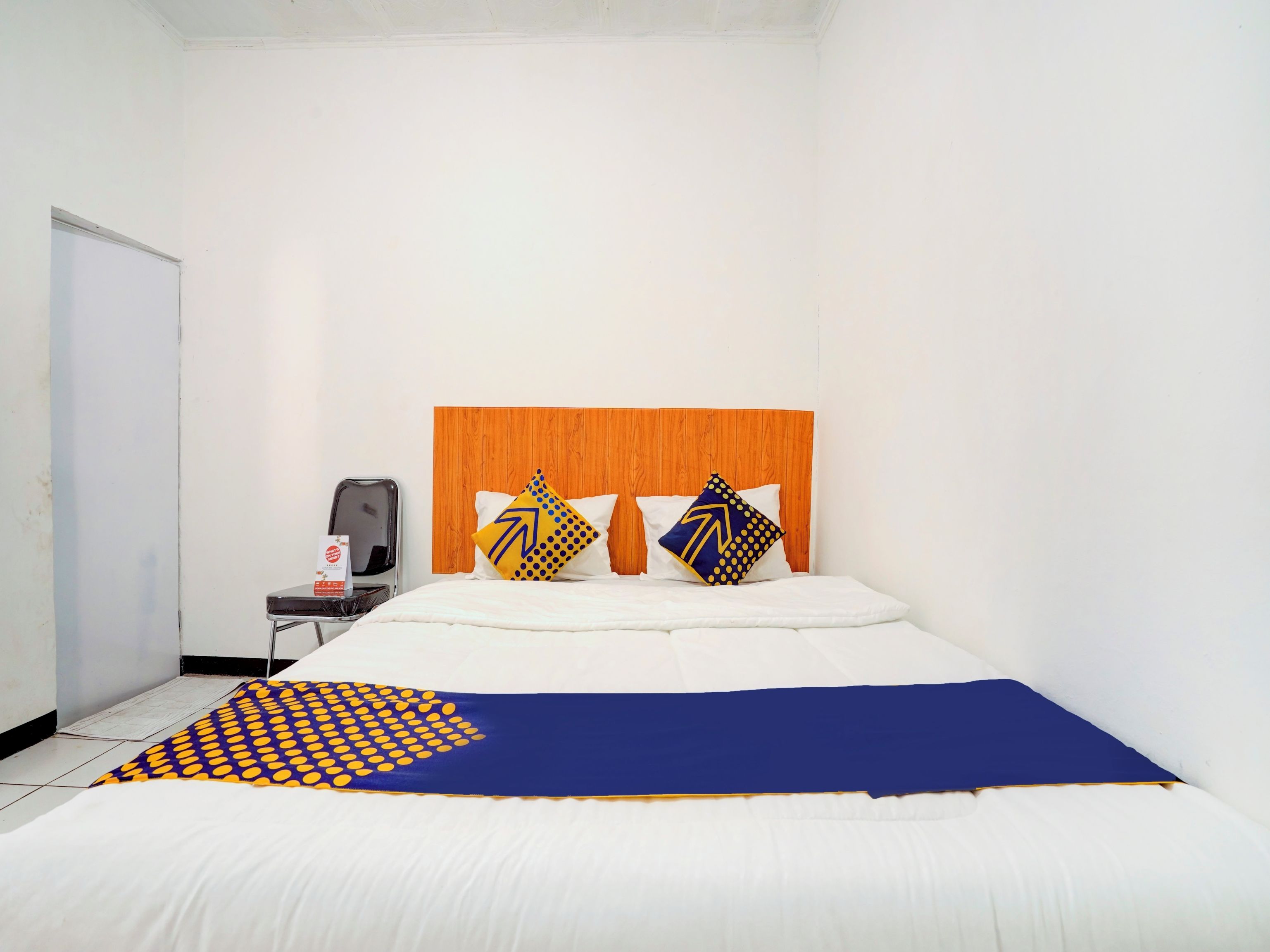 A hotel room with a wooden headboard, a bed with white and blue bedding, two pillows with geometric patterns, a small table with a chair, and a whiteboard on the wall.