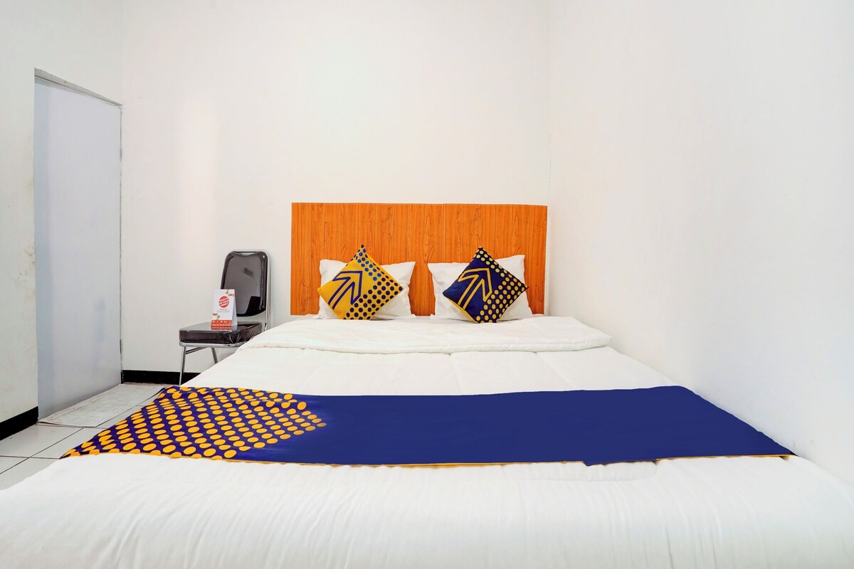 A hotel room with a wooden headboard, a bed with white and blue bedding, two pillows with geometric patterns, a small table with a chair, and a whiteboard on the wall.