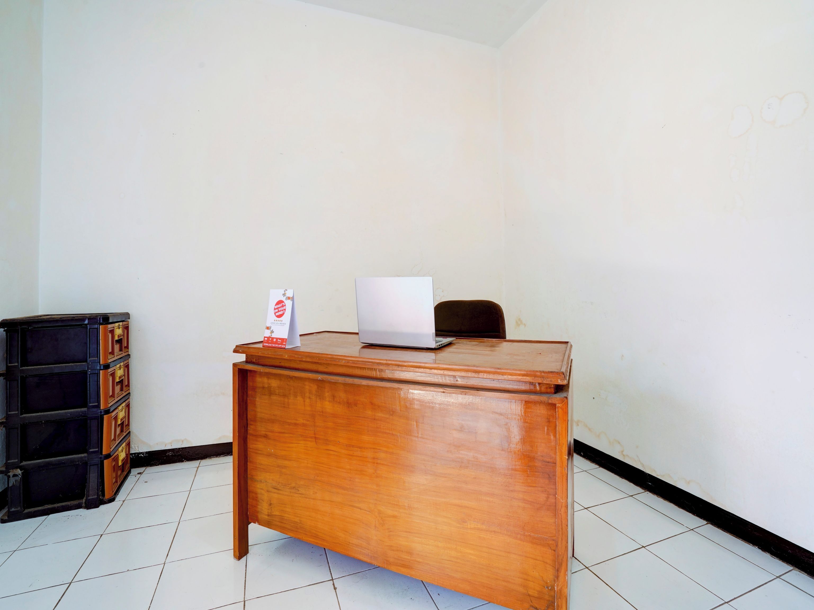 A wooden desk with a laptop and a sign on it, a black filing cabinet, and a tiled floor.
