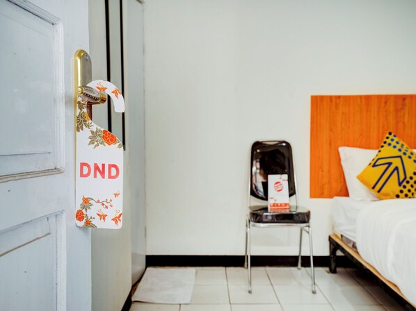 Quarto de hotel com cama, uma cadeira, uma mesinha e porta com placa.