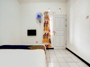 Ein Schlafzimmer mit einem Bett, einem an der Wand befestigten Fernseher, einem Standventilator und einer Tür mit floralem Vorhang.