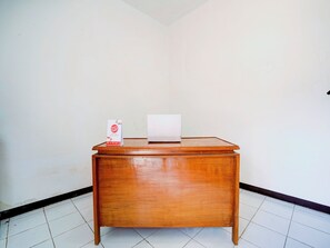 A wooden desk with a laptop and a brochure in a corner of a room with white tiled flooring and plain walls. - Spot ON 90200 Pondok Sabaraya Cileunyi Syariah (Cileunyi)