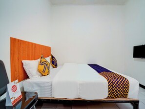 Quarto de hotel com cabeceira de madeira, cama com roupa de cama branca, duas almofadas decorativas, manta com padrão de bolinhas e uma televisão de tela plana fixada na parede.