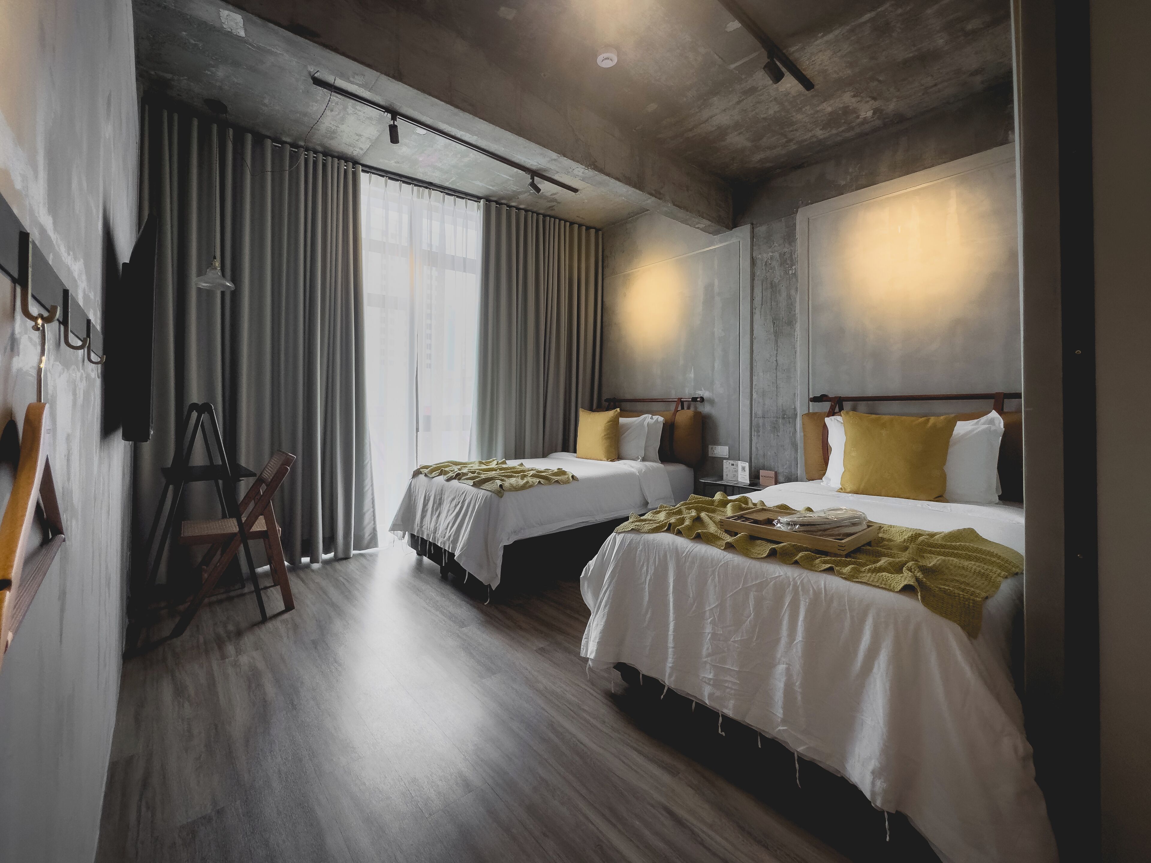 A hotel room with two beds, a wooden floor, a large window with curtains, and a ladder against the wall.