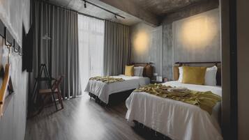 A hotel room with two beds, a wooden floor, a large window with curtains, and a ladder against the wall.