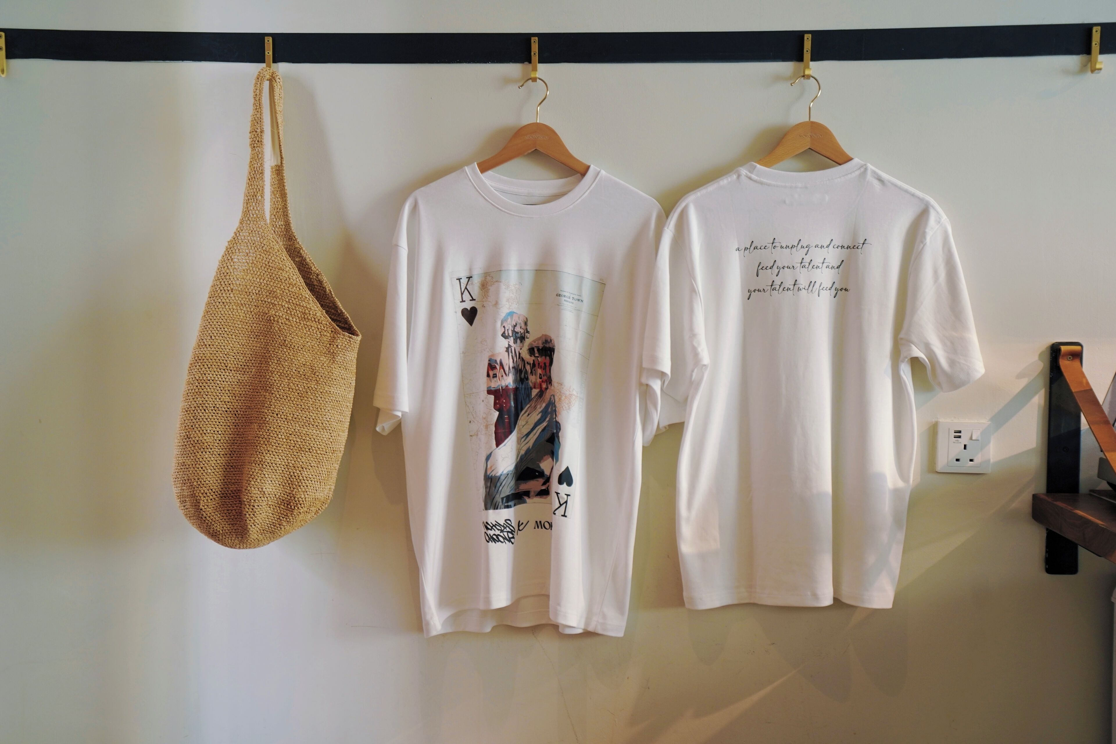 Tres camisetas blancas colgadas en un perchero, una con estampado e inscripción, y dos lisas con texto en la parte posterior.