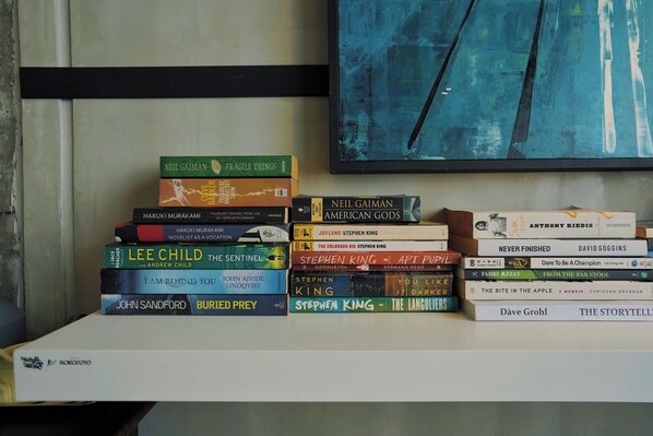 A stack of books on a white shelf against a wall with a blue abstract painting.