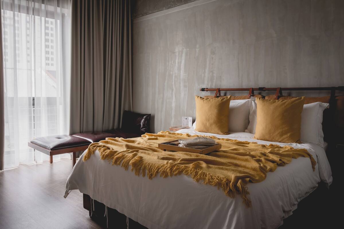 A hotel room with a large bed, a wooden bench, and a view of buildings through sheer curtains.