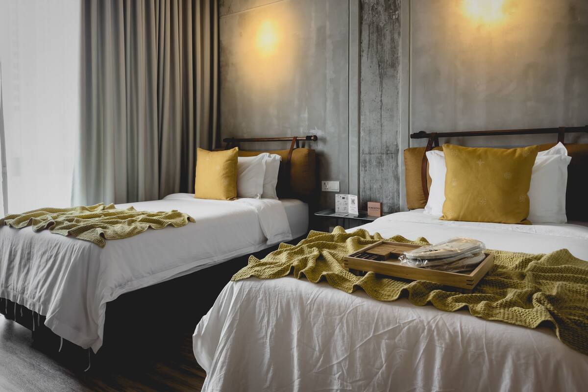 A hotel room with two beds, each with white linens and yellow pillows, a wooden nightstand with a lamp, and a window with curtains.