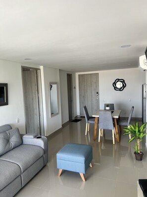Een moderne woonkamer met een grijze bank, een blauwe poef en een eethoek met grijze stoelen en een kleine tafel.