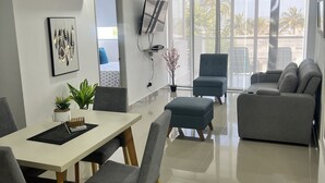 Un salon moderne comprenant un coin repas, des canapés gris, un téléviseur à écran plat et une grande fenêtre donnant sur des palmiers.