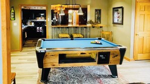 A pool table in a room with wooden beams, a patterned rug, and a kitchen in the background. - Limestone Corner Bungalow (Graytown)