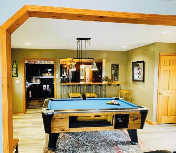 A pool table in a room with wooden beams, a patterned rug, and a kitchen in the background. - Limestone Corner Bungalow (Graytown)