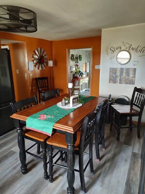 A dining room with a wooden table, chairs, and a green table runner. There is a wall with a 'Stay Awake' sign and a decorative mirror. - Limestone Corner Bungalow (Graytown)