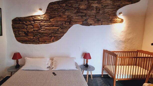 Une chambre avec un mur en pierre, un lit avec une literie blanche, deux lampes rouges et un berceau en bois.