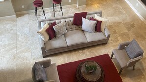 Un vaste espace de vie comprenant un canapé, deux fauteuils, une table basse et un tapis rouge. On aperçoit, au fond, une cuisine équipée d’appareils électroménagers modernes.