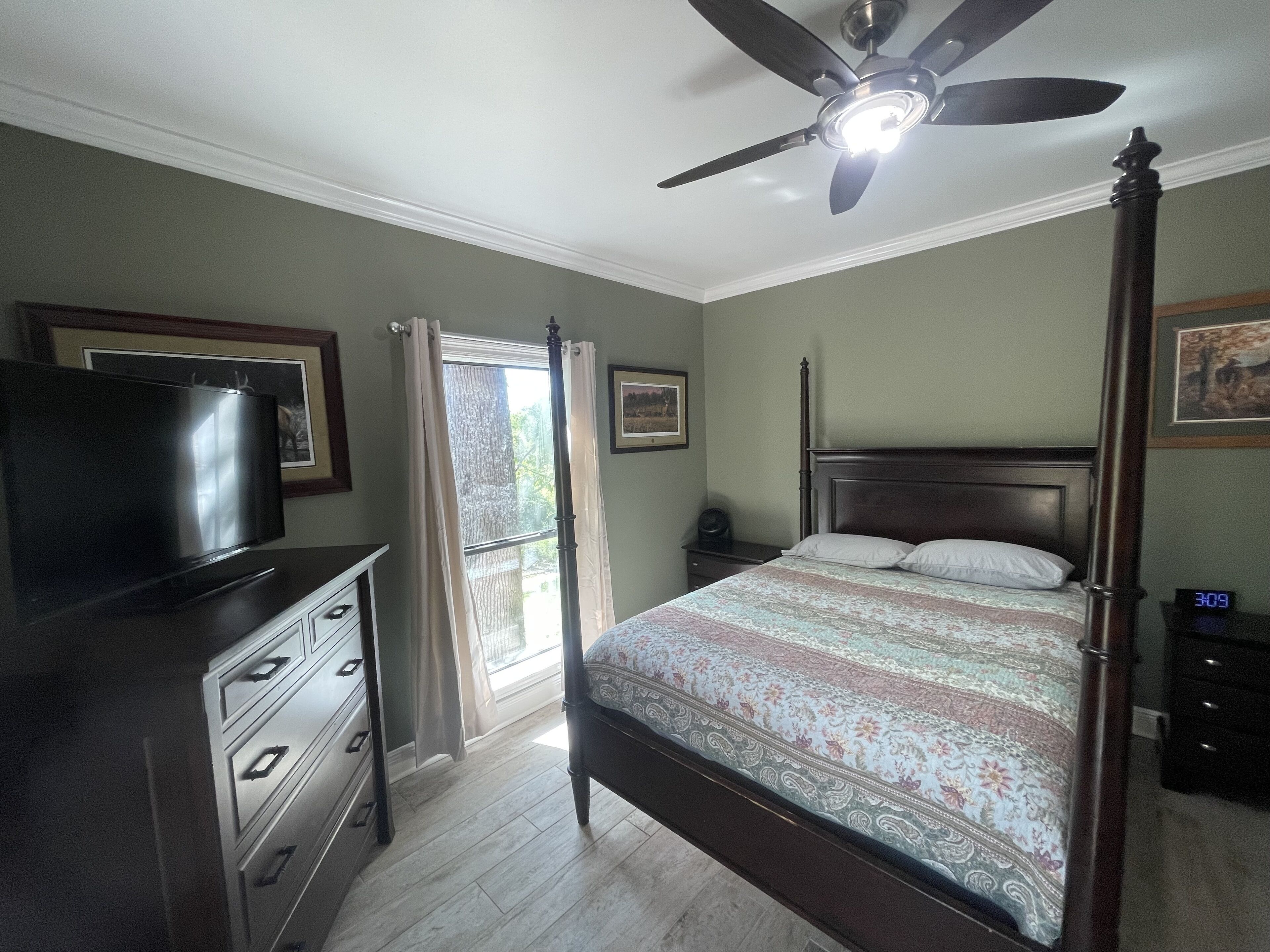 Un dormitorio con cama, televisor, cómoda, mesita de noche, ventilador de techo y ventana con cortinas.