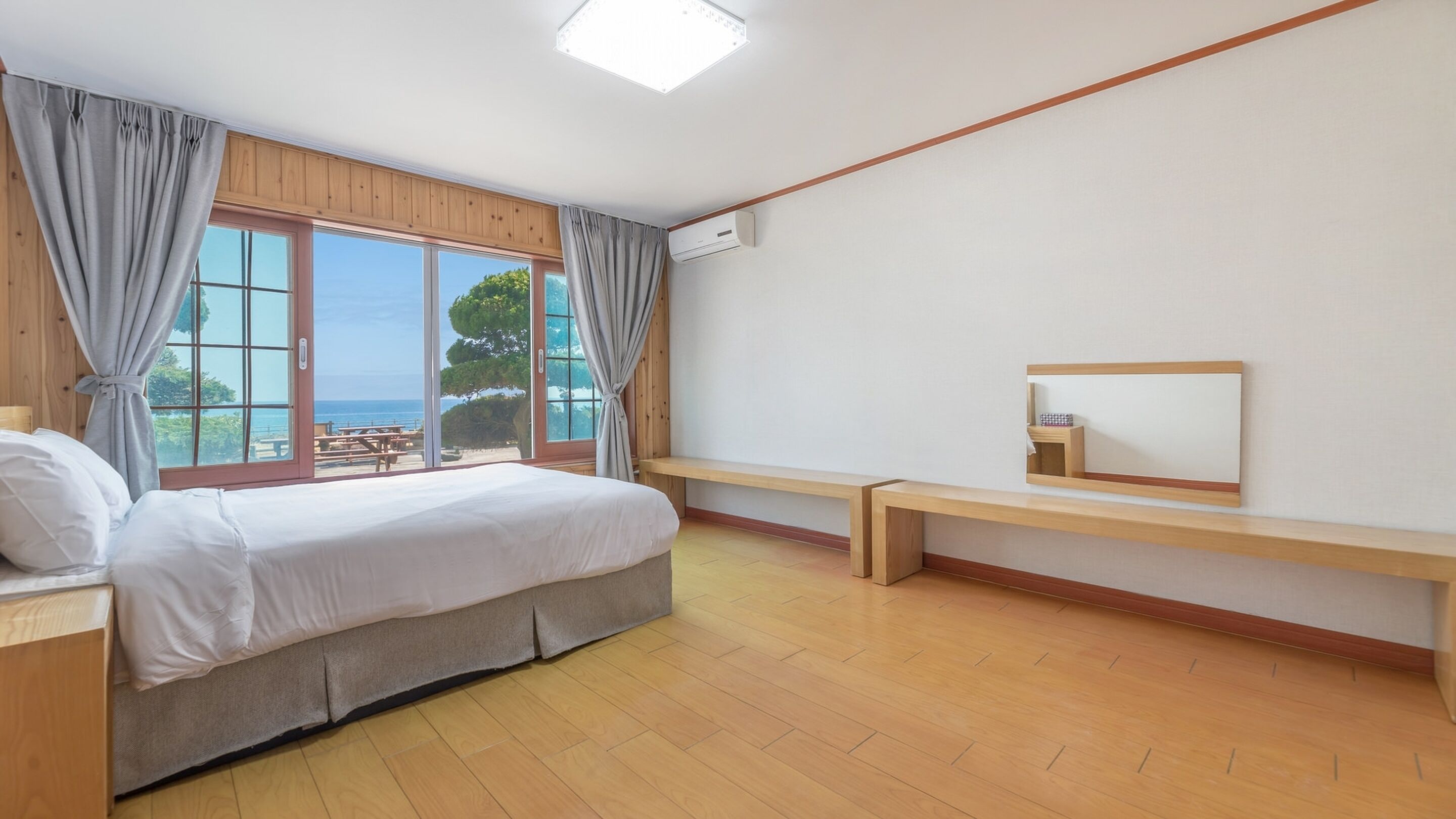 A bedroom with a large window, a bed, a bench, and a mirror.