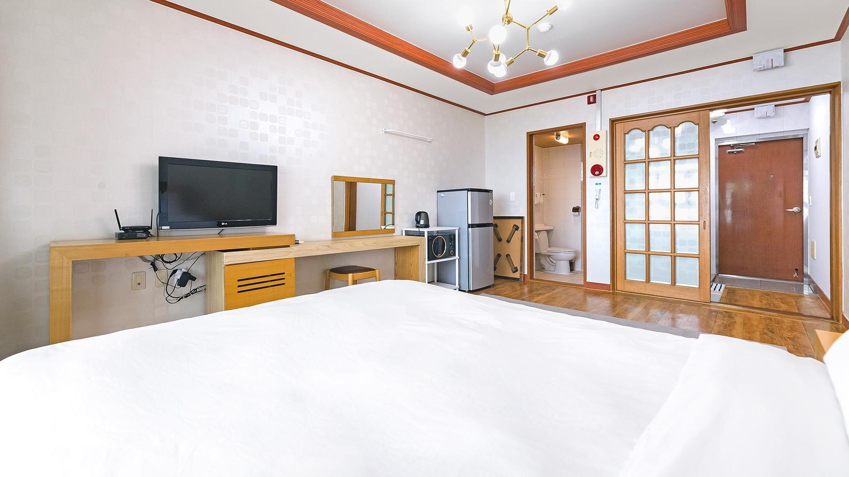 A hotel room with a bed, a desk with a TV, a microwave, and a refrigerator. There is a door leading to a bathroom with a toilet and a shower.