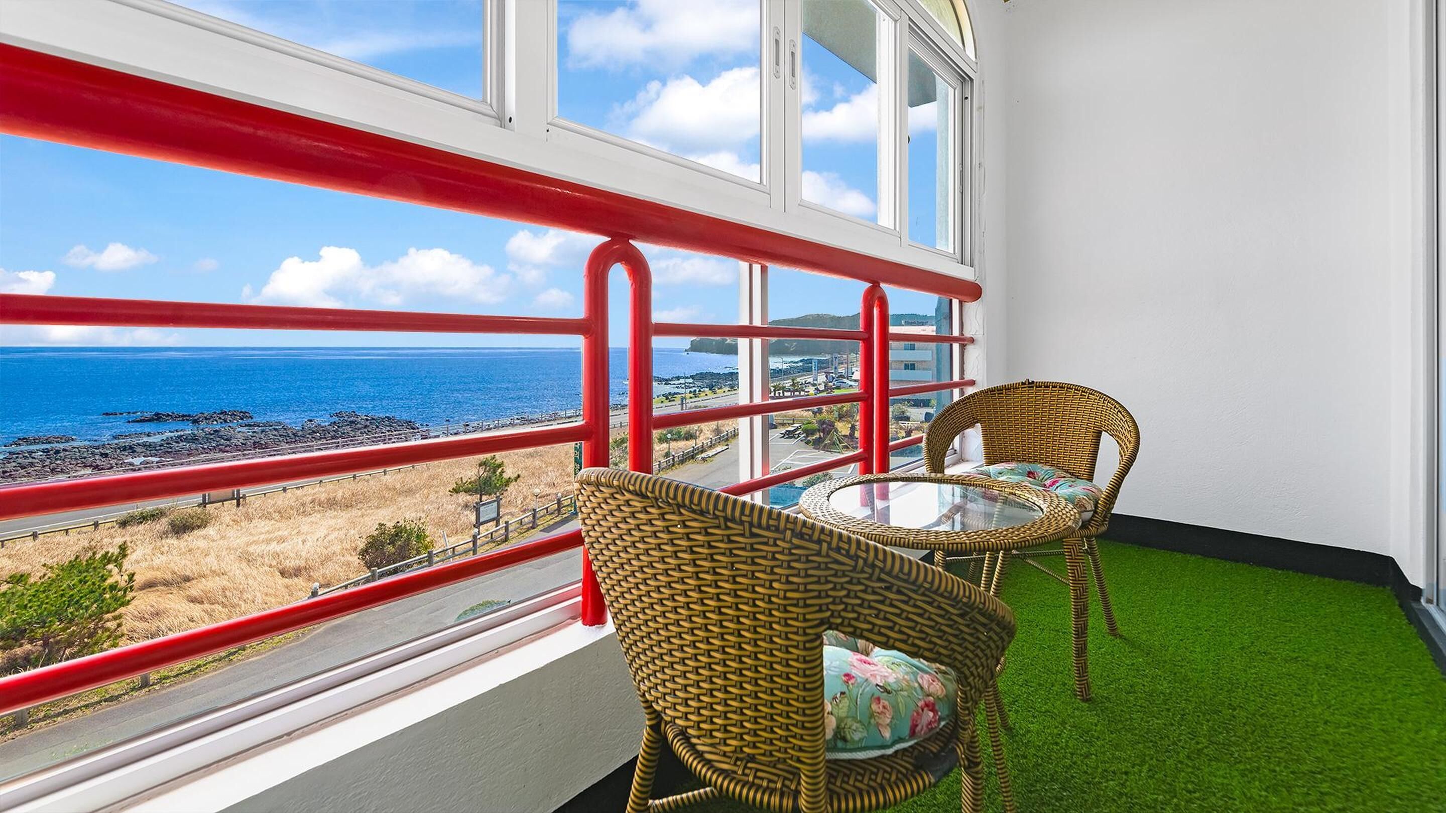 Un balcón con sillas de mimbre y una mesa de vidrio, con vista al mar y una barandilla roja.