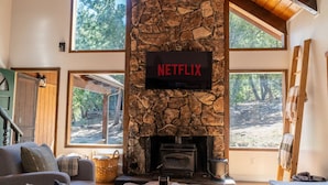 Ein gemütliches Wohnzimmer mit Kamin aus Stein, einem Netflix-Schild, großen Fenstern und einer bequemen Couch.