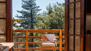 A wooden deck with a chair and a table, leading to a balcony with a view of trees. - Secluded 1 acre Mountain Home - Hot Tub, 3 Decks & Direct National Forest Access (Idyllwild-Pine Cove)